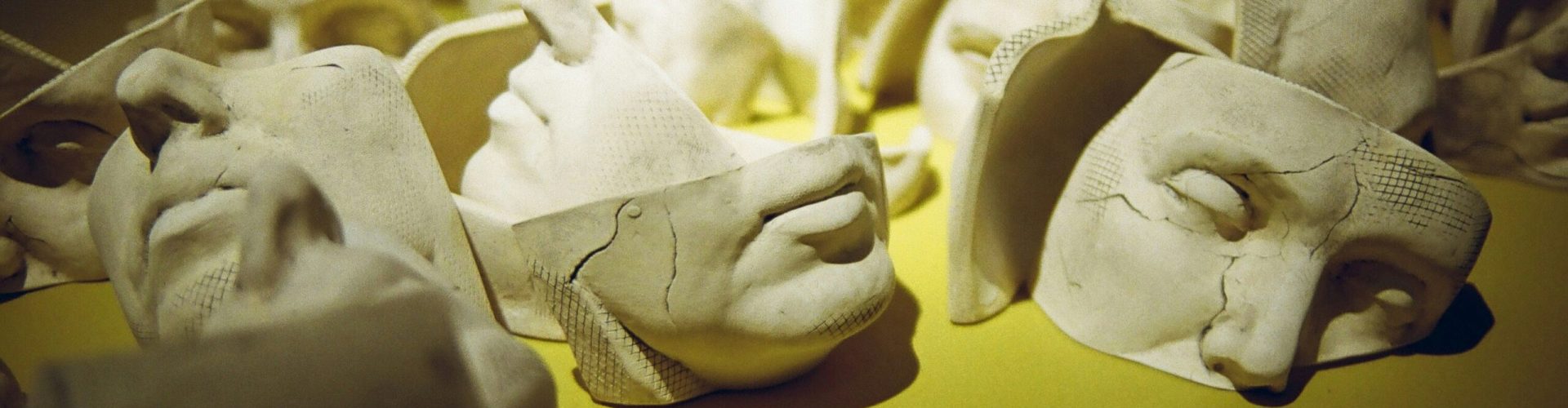 Mehrere zerbrochene Gipsmasken liegen auf einer gelben Oberfläche, die Fragmente zeigen Gesichter mit feinen Rissen und Bruchstellen.