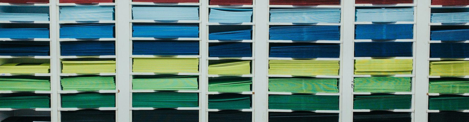 shawn-r2A6WYI8YIg-unsplash Regale mit vielen Fächern, gefüllt mit farbig sortierten Papierstapeln. Die Farben reichen von Gelb, Orange, Rosa und Rot über Blau und Grün bis hin zu Weiß und Schwarz. Die Papiere sind ordentlich gestapelt und in vertikalen Reihen angeordnet, was ein buntes, strukturiertes Muster ergibt.