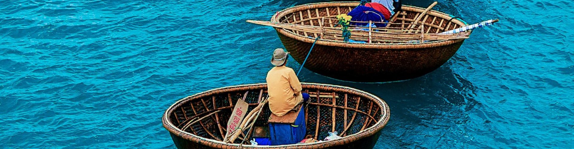 Mehrere runde, geflochtene Korbboote mit sitzenden Personen auf türkisblauem Wasser hintereinander angeordnet.